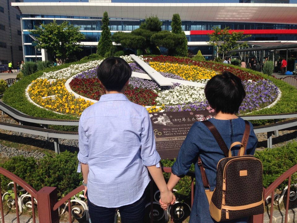 무밍+K 커플이 꽃시계 앞에 손을 잡고 있는 모습.